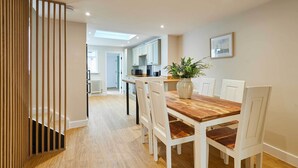 A dining area with a wooden table and white chairs, a potted plant, and a view into the kitchen. - Host & Stay | Ginger Bread Cottage (Stokesley)