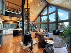 A spacious interior with a high ceiling, wooden floors, a central seating area with a sofa and armchairs, a kitchen area, and large windows offering a view of the outdoors.