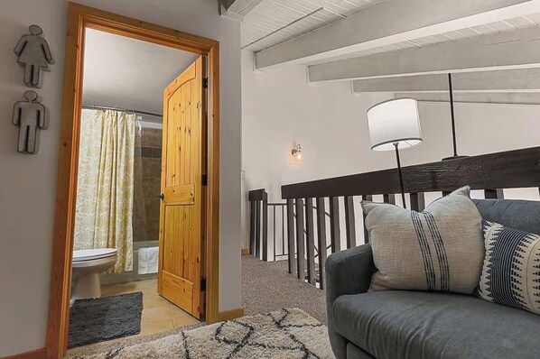 A living room with a grey sofa, patterned cushions, a wooden door leading to a bathroom, and a staircase with a railing.