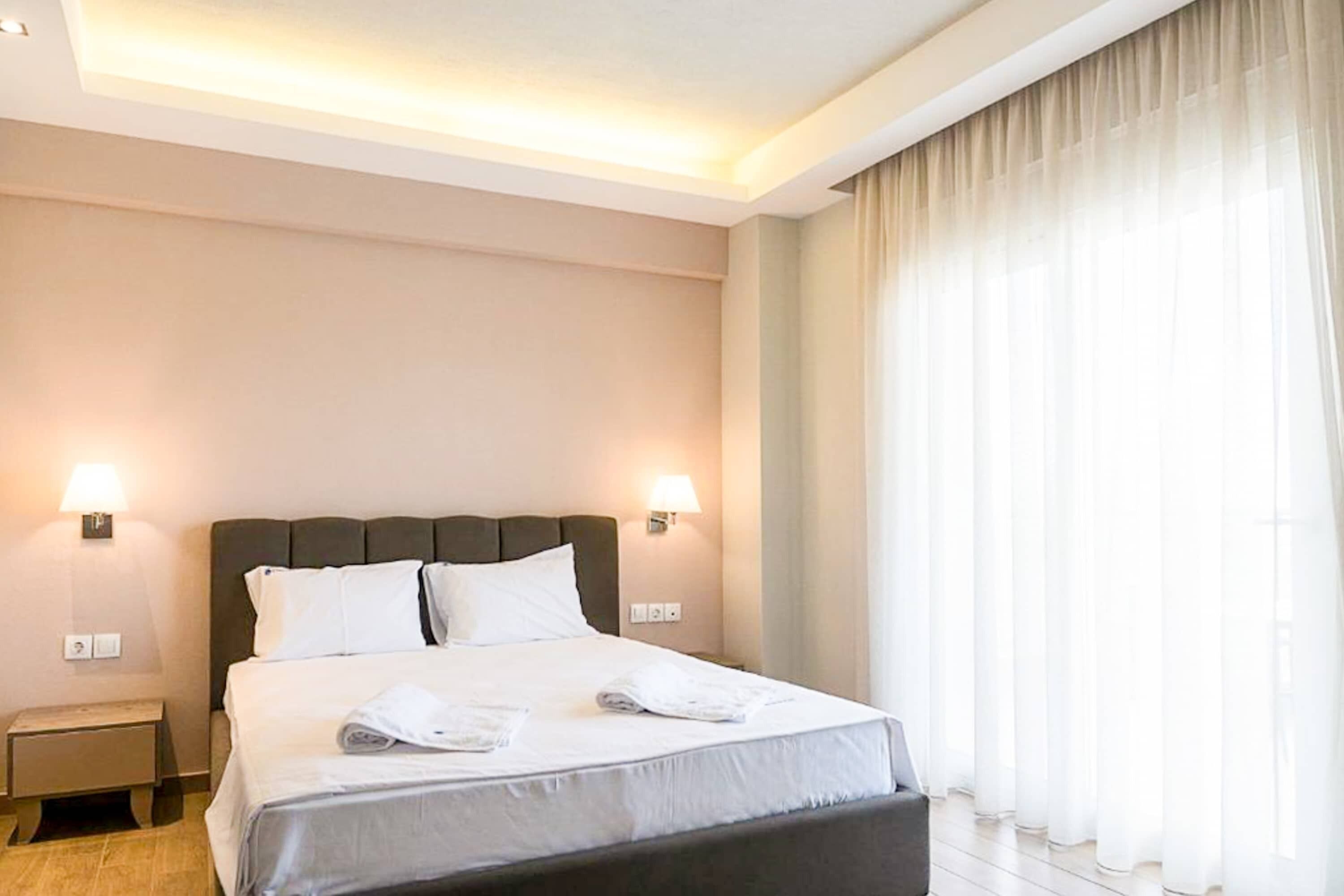 A hotel room with a large bed, two bedside lamps, a nightstand, and sheer curtains.