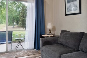 A living room with a sofa, a small table with a lamp, and a view of greenery through a window.