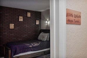 Ein Einzelbett mit einem gemusterten Bettbezug in einem Zimmer mit Backsteinwand und einem Schild mit der Aufschrift 'Wizards Welcome - Muggles Tolerated'.