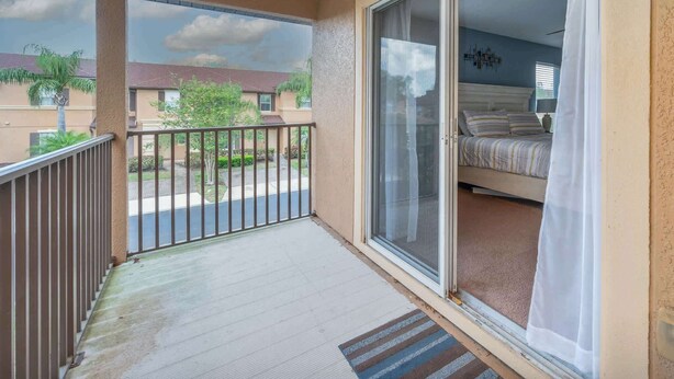 Uma varanda com vista para as casas vizinhas, uma porta de correr de vidro que dá para um quarto com cama e cortinas, e um tapete na entrada.