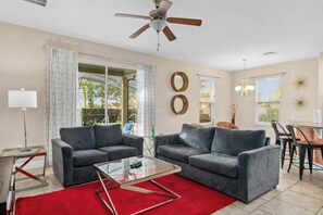 A living room with two grey sofas, a glass coffee table, a red rug, a ceiling fan, and a dining area with a table and chairs.