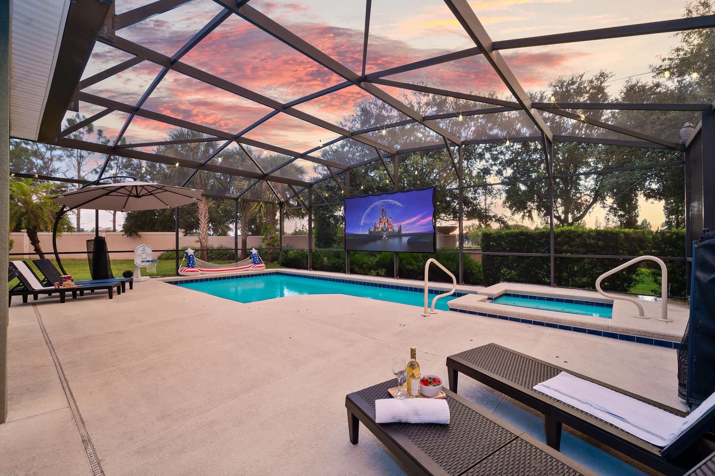A covered outdoor pool area with lounge chairs, a table with a bottle and glass, and a TV screen displaying a castle.