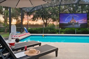 A poolside area with lounge chairs, a large screen displaying a castle, and a view of a sunset. - Resort Villa w/ Private Pool & Family Fun (Davenport)