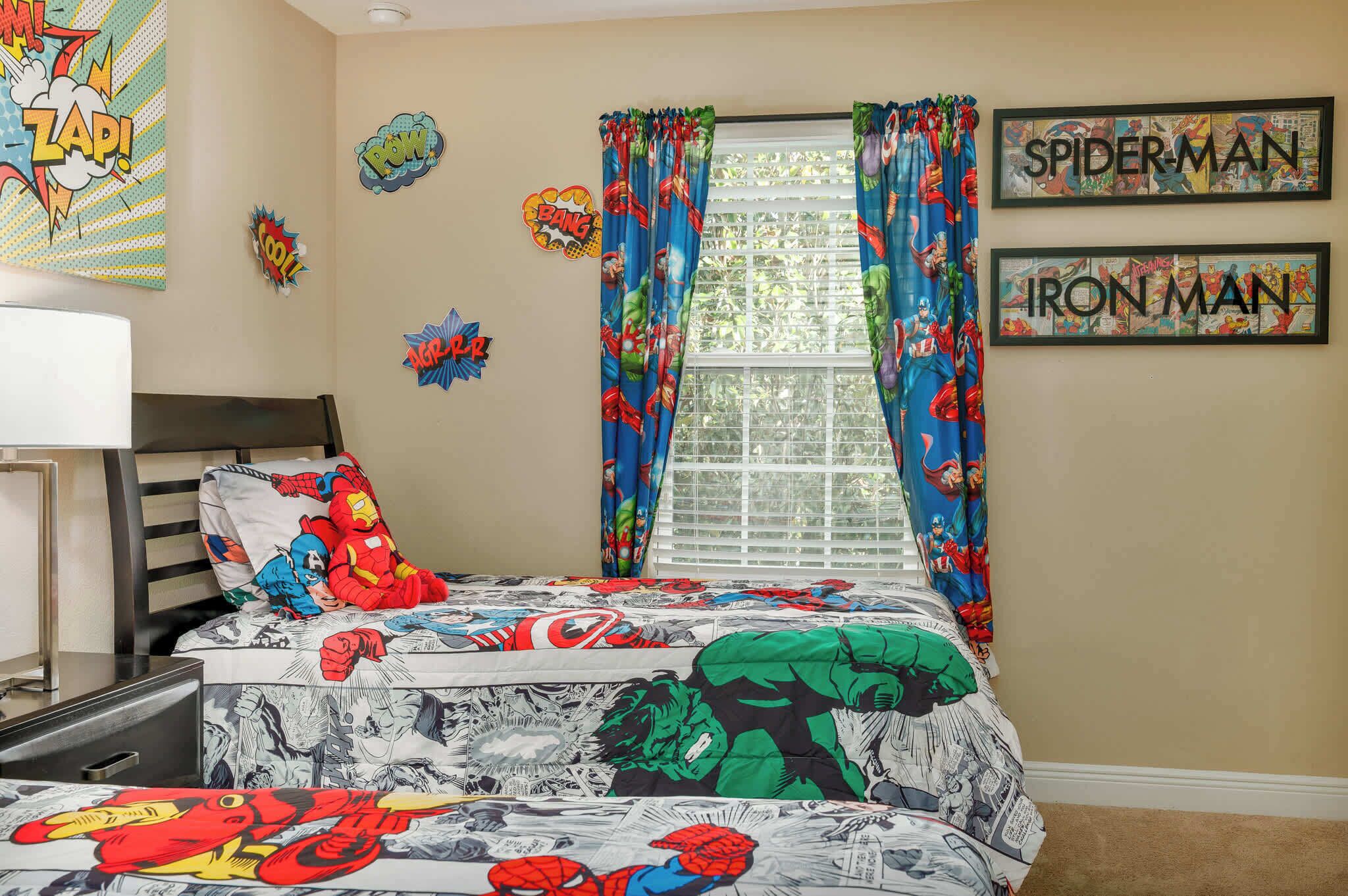A bedroom with a bed featuring superhero-themed bedding, a window with curtains, and superhero wall art.