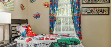 A bedroom with a bed featuring superhero-themed bedding, a window with curtains, and superhero wall art.