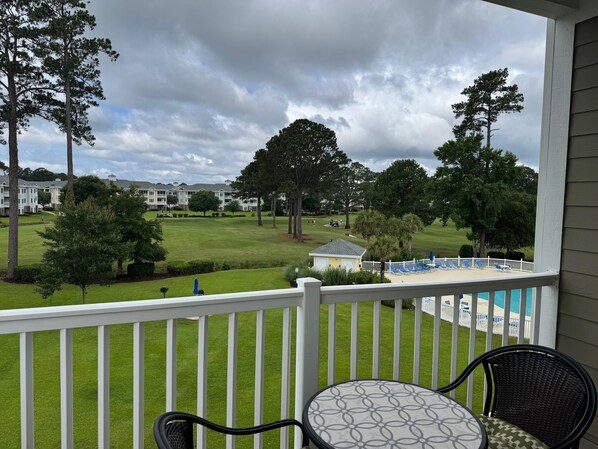 Un balcón con vistas a un campo de golf, una zona de piscina con sillones de descanso y un edificio pequeño a lo lejos.