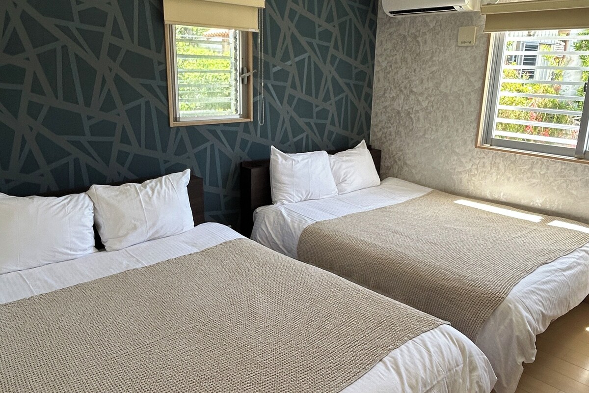 A hotel room with two beds, a window with blinds, a patterned wall, and an air conditioning unit.