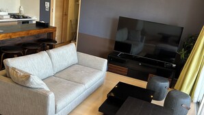 Un salon moderne avec un canapé gris, un pouf et une table basse noire, un téléviseur à écran plat et un coin cuisine équipé d’une cuisinière et de tabourets de bar.