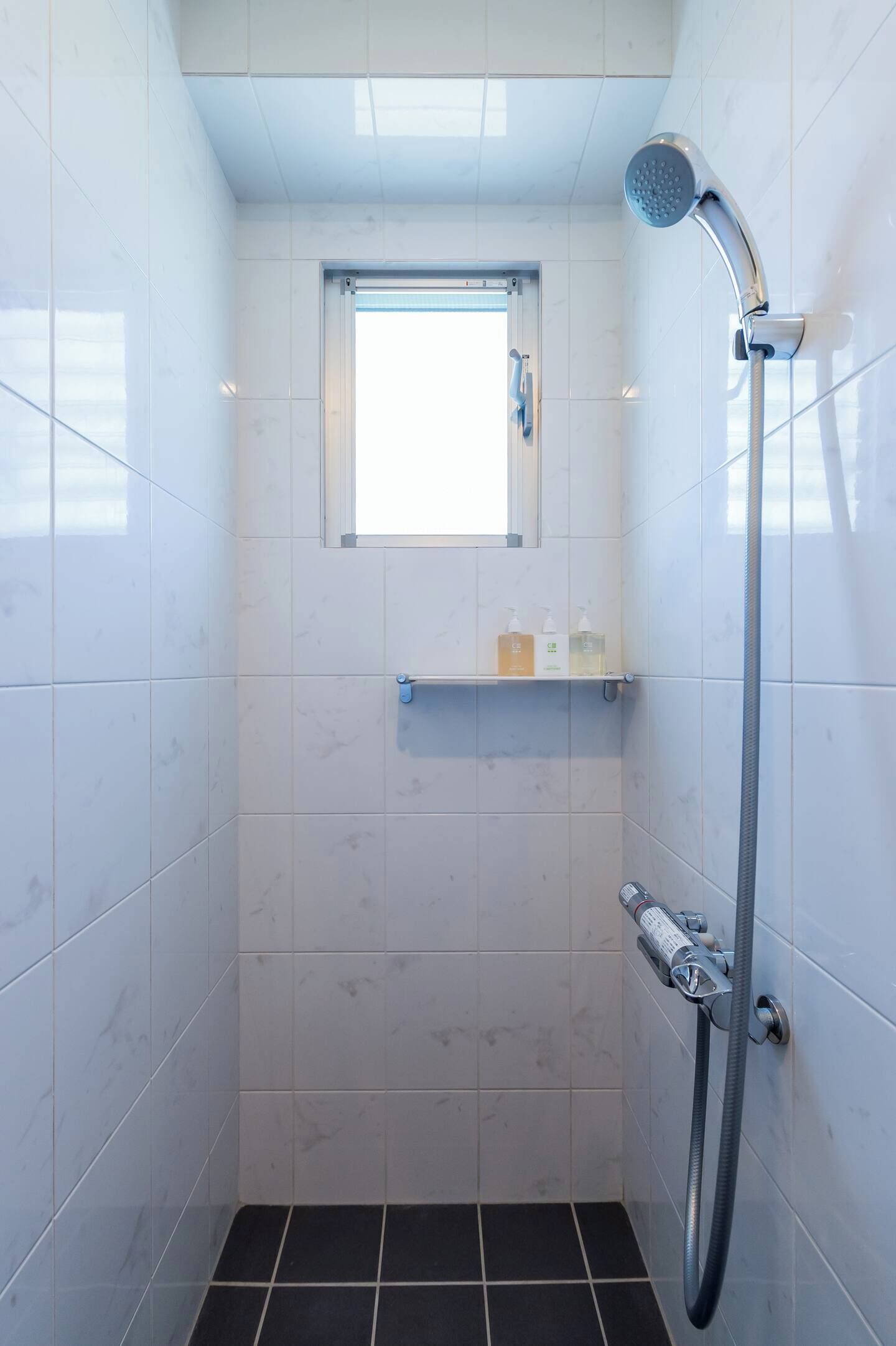 A small, modern bathroom with white tiles, a showerhead, a window, and a shelf with toiletries.