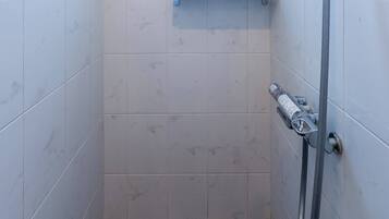 A small, modern bathroom with white tiles, a showerhead, a window, and a shelf with toiletries.