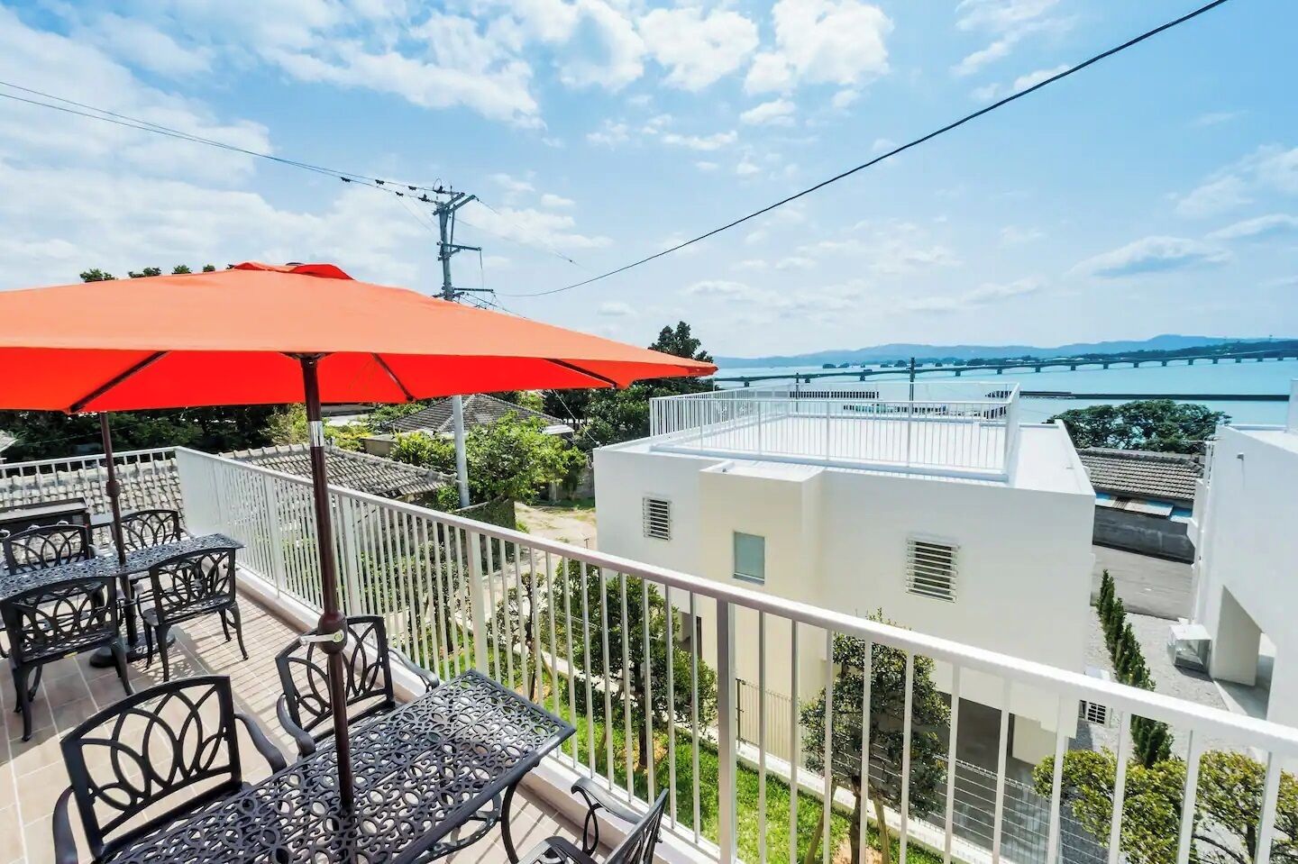 A balcony with outdoor furniture and a red umbrella, overlooking a cityscape with a bridge and water body.