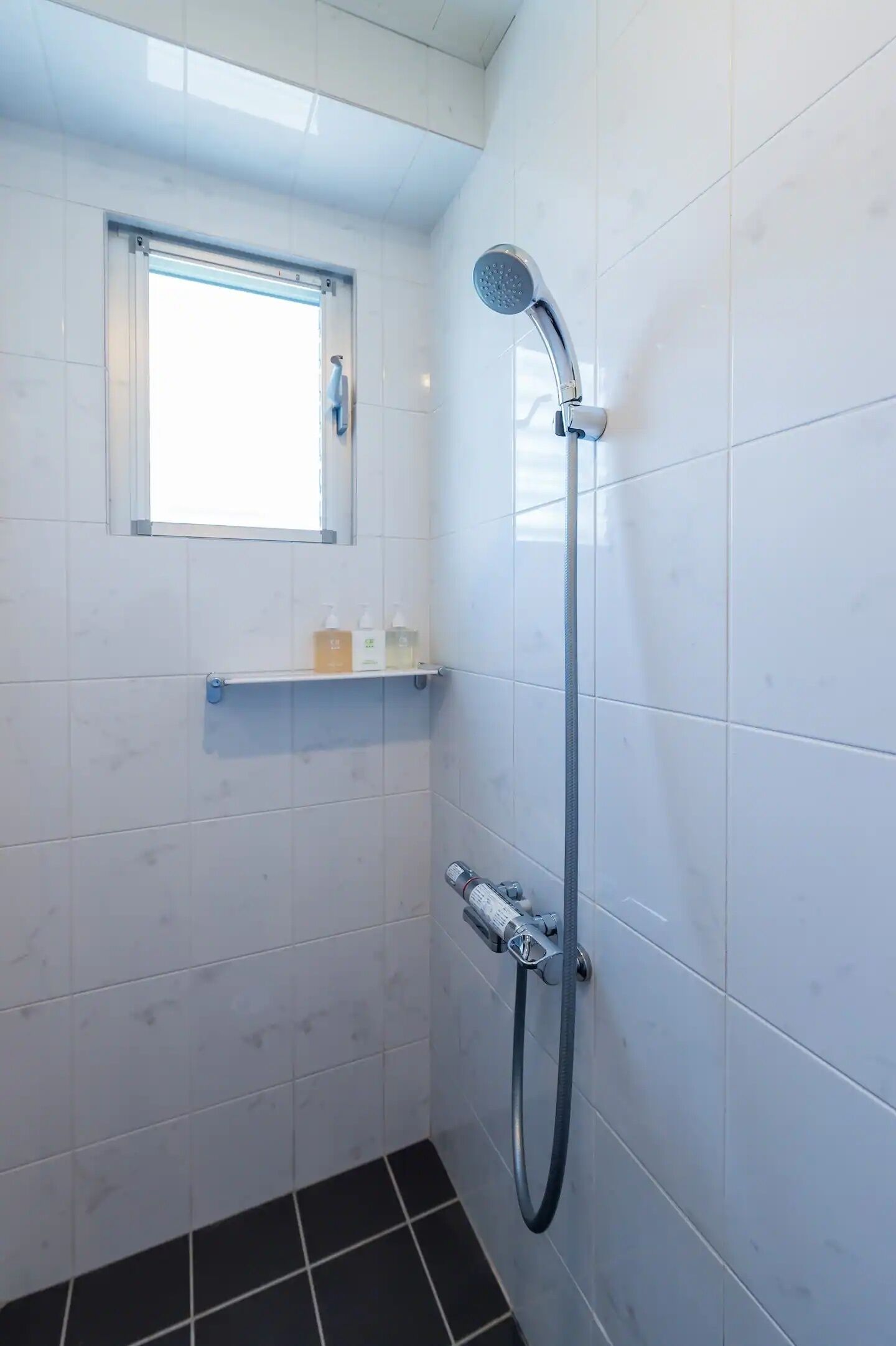 A small bathroom with white tiled walls, a showerhead, a window, and a shelf with toiletries.
