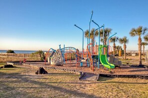 Ein Spielplatz mit verschiedenen Spielgeräten, darunter eine Rutsche und Klettergerüste, umgeben von einem eingezäunten Bereich mit Palmen.