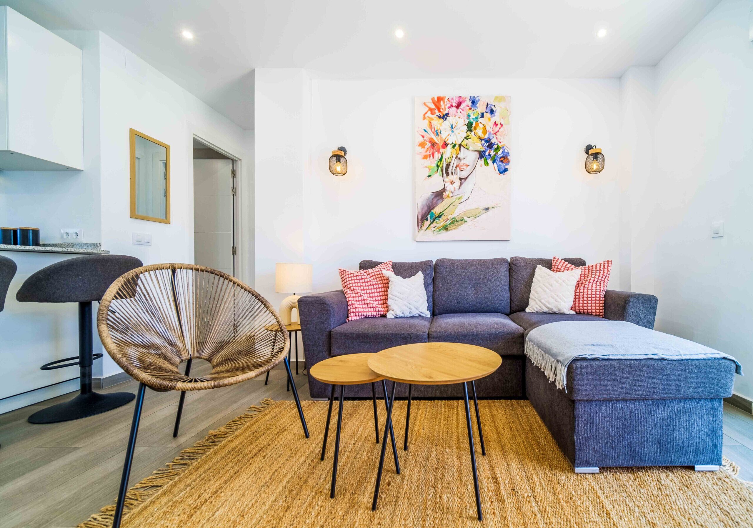 A modern living room with a sofa, two small round tables, a wicker chair, and a painting on the wall.