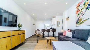 Een moderne woonkamer met een grijze bank, houten kasten, een televisie en een keuken op de achtergrond.