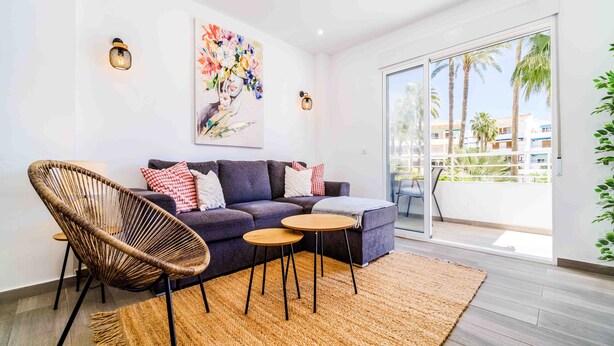 Een moderne woonkamer met een bank, twee ronde tafels, een rieten stoel en een groot raam met uitzicht op palmbomen en gebouwen.