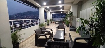 Un balcón con mobiliario de mimbre, plantas en macetas y vista a la ciudad al anochecer.