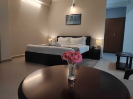 Una habitación de hotel con cama, mesitas de noche, una mesa redonda con un florero y un cuadro en la pared.