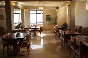 Un comedor amplio con varias mesas y sillas de madera, ventanas grandes que dejan entrar luz natural, y lámparas fijadas a la pared.