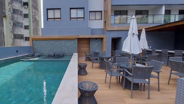 Un área de piscina en la azotea con una piscina azul cristalina, rodeada de sillas y mesas de mimbre, y un edificio moderno al fondo.