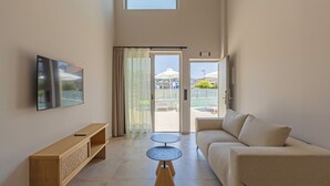 Una sala moderna con una televisión de pantalla plana, una mesa consola de madera, un sofá color beige y una mesa de centro de vidrio. Hay una puerta corrediza de vidrio que da a un área exterior con sombrillas y una piscina.