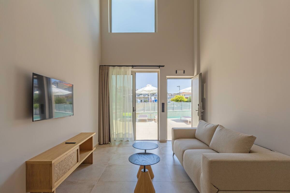 Una sala moderna con una televisión de pantalla plana, una mesa consola de madera, un sofá color beige y una mesa de centro de vidrio. Hay una puerta corrediza de vidrio que da a un área exterior con sombrillas y una piscina.