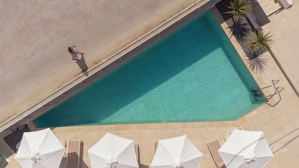 Vista aérea de una piscina con solárium azulejado, rodeada de sombrillas blancas y sillones de descanso.