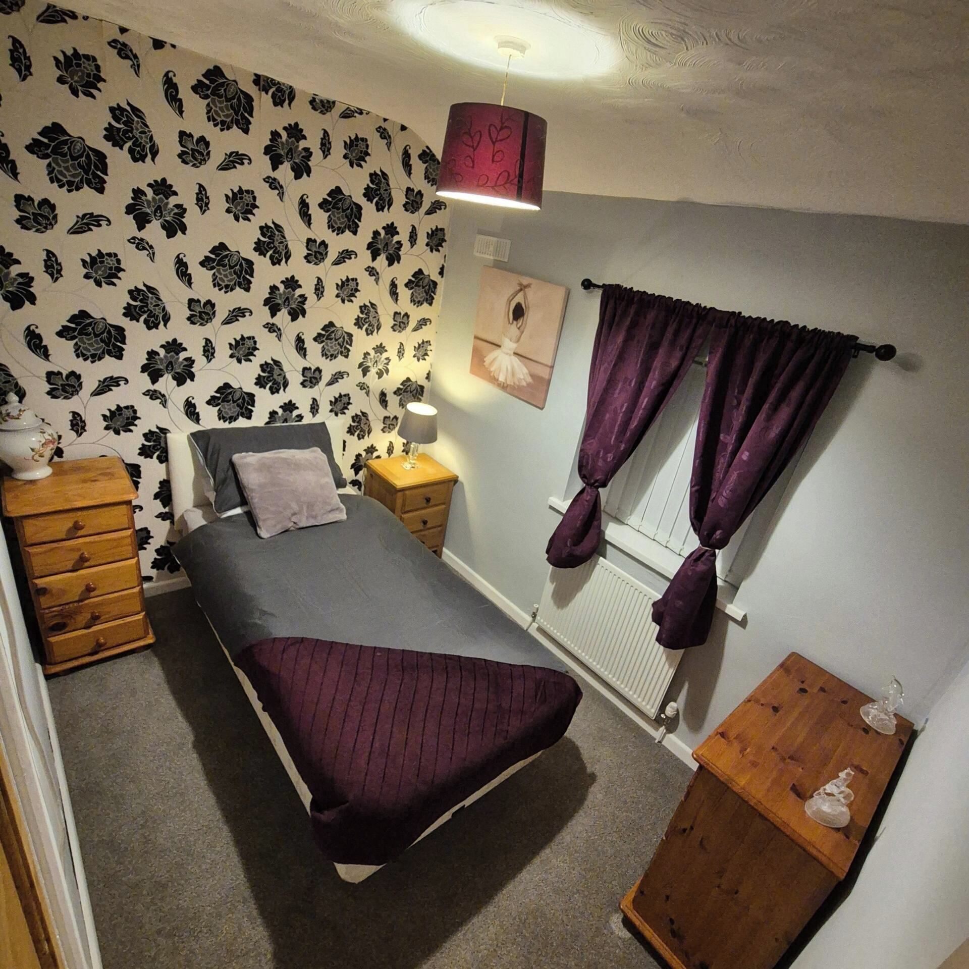 A bedroom with a bed, a dresser, a nightstand, a lamp, a window with curtains, and a radiator.