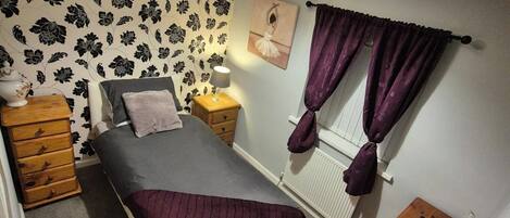 A bedroom with a bed, a dresser, a nightstand, a lamp, a window with curtains, and a radiator.