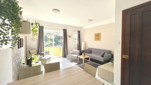 Een moderne woonkamer met een bank, salontafel en eethoek. Er is een groot raam met gordijnen, een plant en een deur.