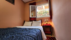 A bedroom with a bed, a nightstand with a lamp, a window with a view of the outside, and a mirror on the wall.