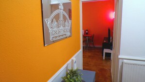 Een kamer met een oranje muur, een spiegel die een kroonontwerp weerspiegelt, een klein tafeltje met een plant en een rode deur naar een andere kamer.