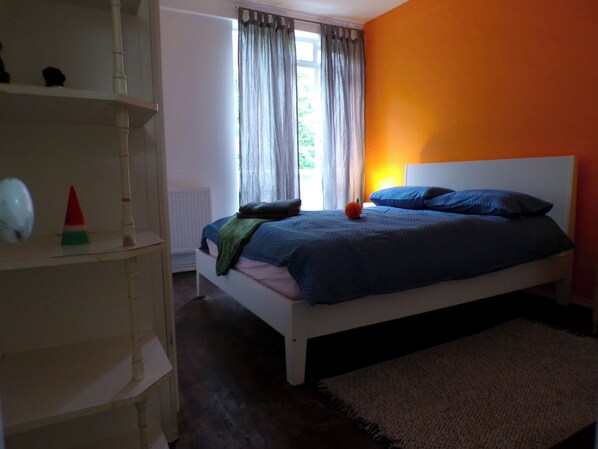 Une chambre avec un lit blanc, une literie bleue, une table de chevet en bois, un radiateur blanc et une fenêtre avec des rideaux.
