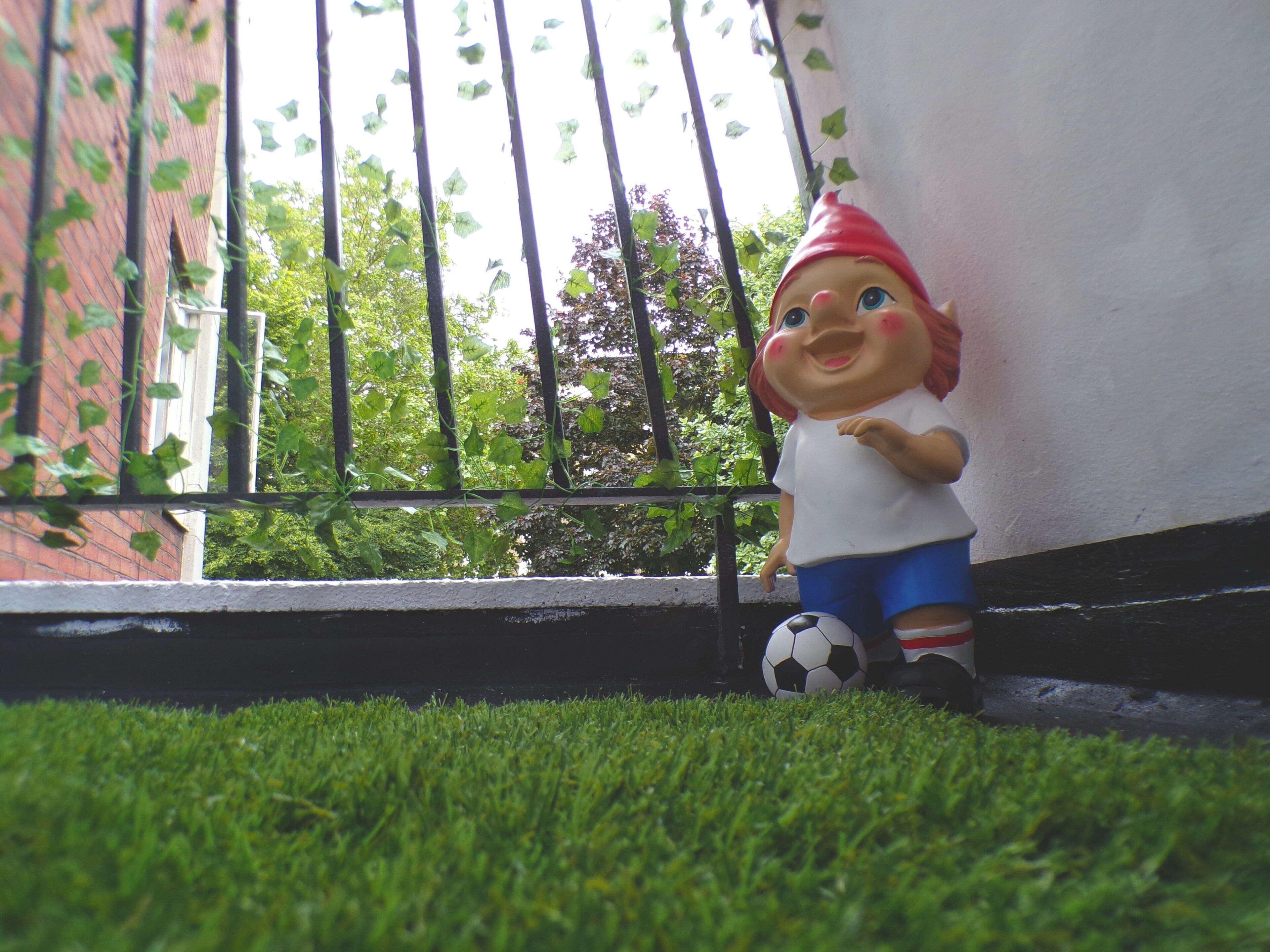Une petite figurine représentant un enfant avec un ballon de football, posée sur une surface herbeuse près d’un bâtiment.