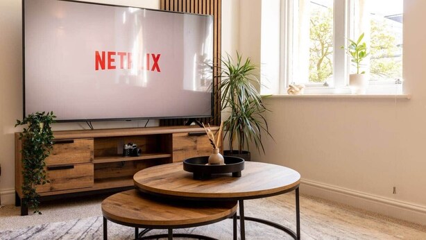 A living room with a large flat-screen TV displaying the Netflix logo, a wooden coffee table, a potted plant, and a wooden cabinet with drawers.