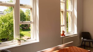 A room with large windows, a radiator, a chair, and a bed with an orange quilt.