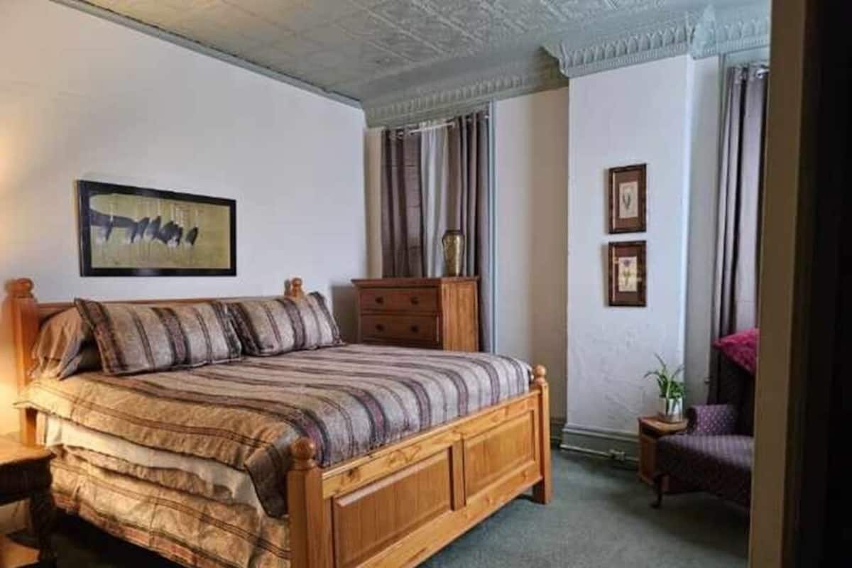 A bedroom with a wooden bed, a nightstand, a dresser, a framed picture on the wall, and a window with curtains.