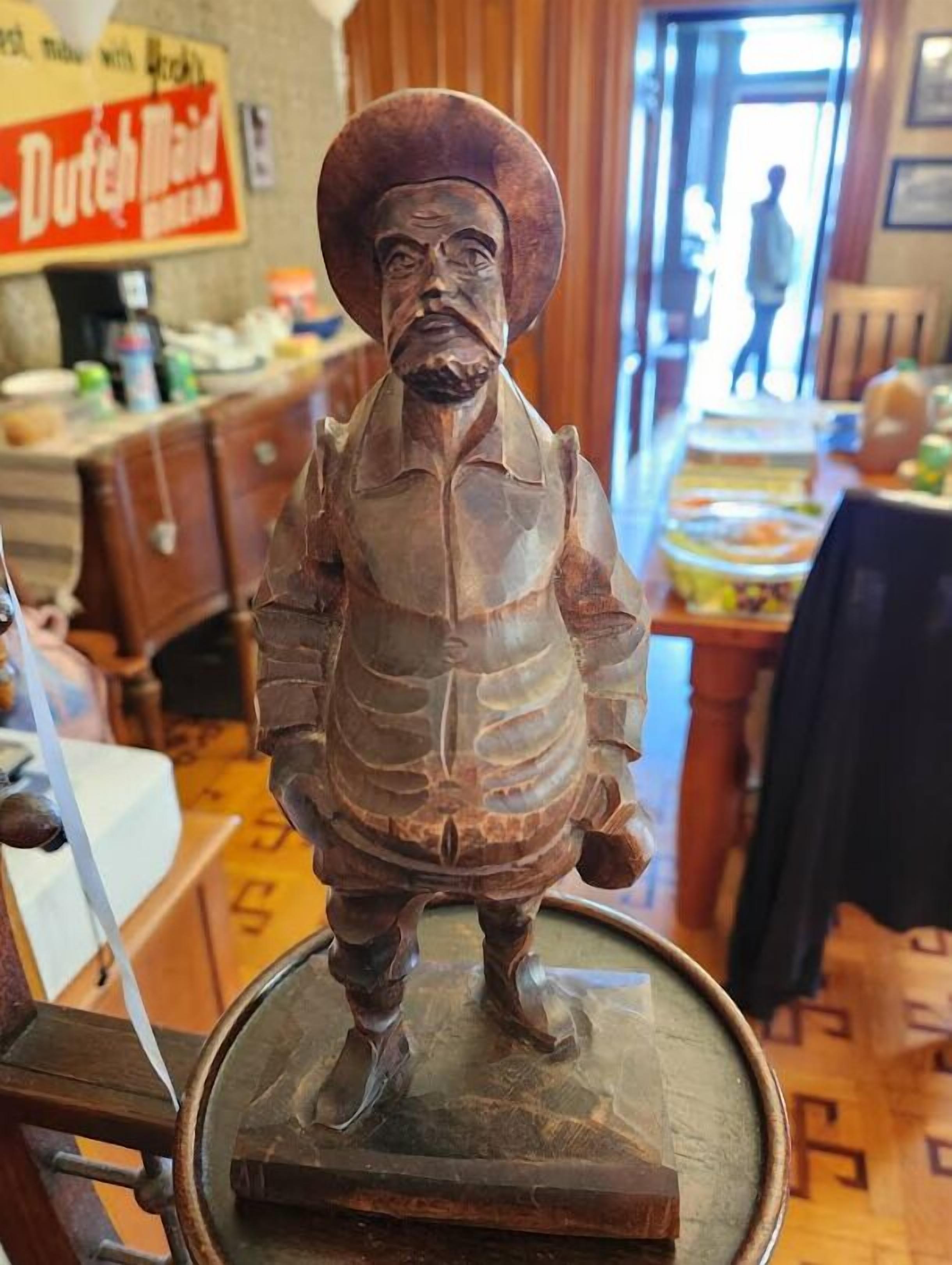 Uma escultura de madeira de um homem com chapéu e colete, colocada sobre uma mesa em um quarto com mobiliário de madeira e uma placa com a inscrição 'Dutch Maid'.