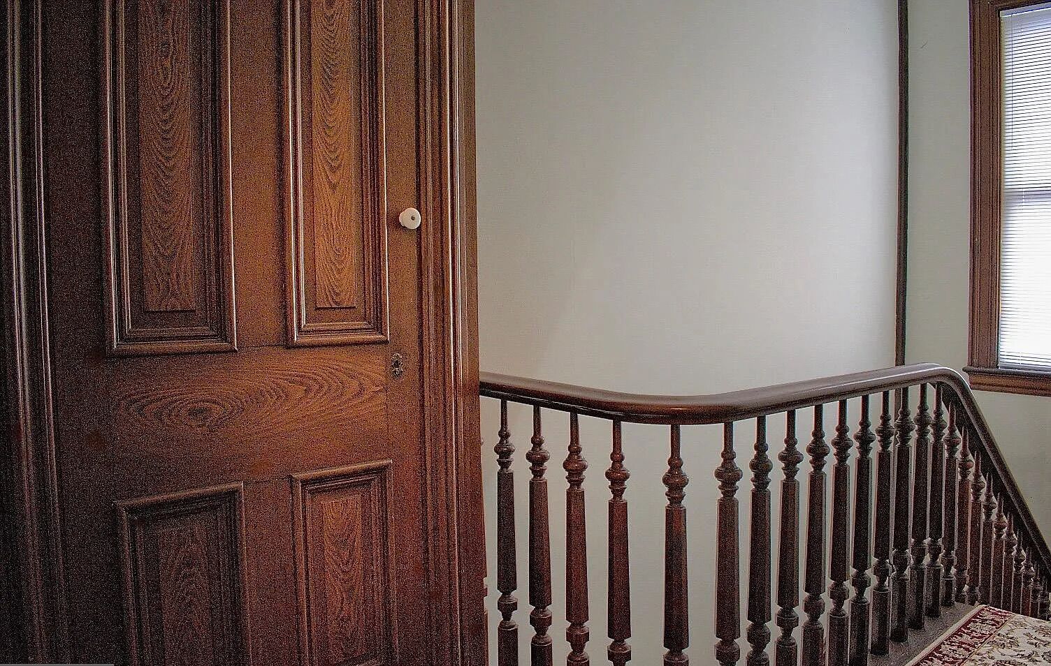 Eine Holztür mit Messingschlussel, eine Holztreppe mit Geländer und ein Fenster mit Jalousien.