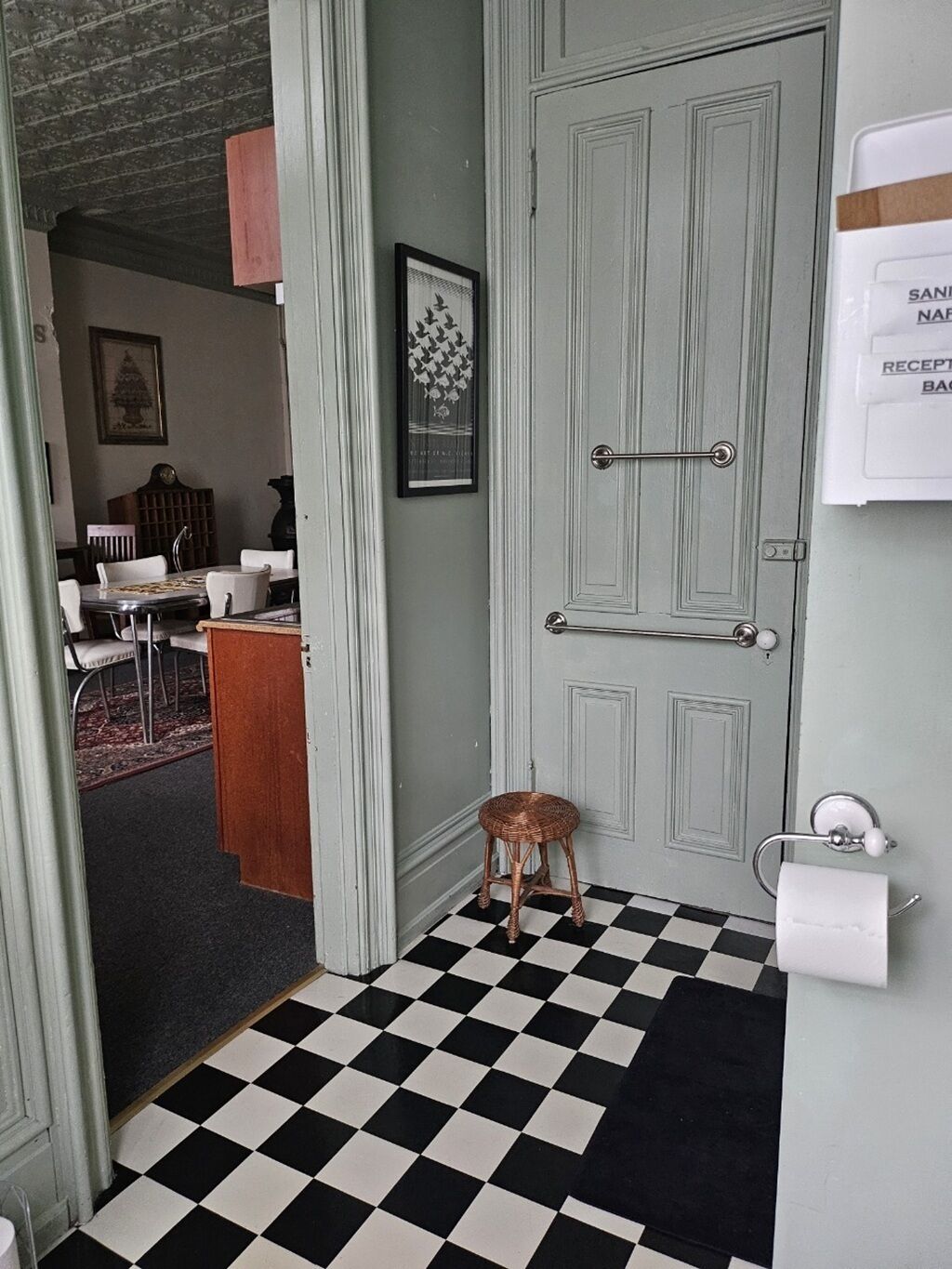 Un baño con piso a cuadros, un taburete pequeño, un perchero y un cuadro en la pared.
