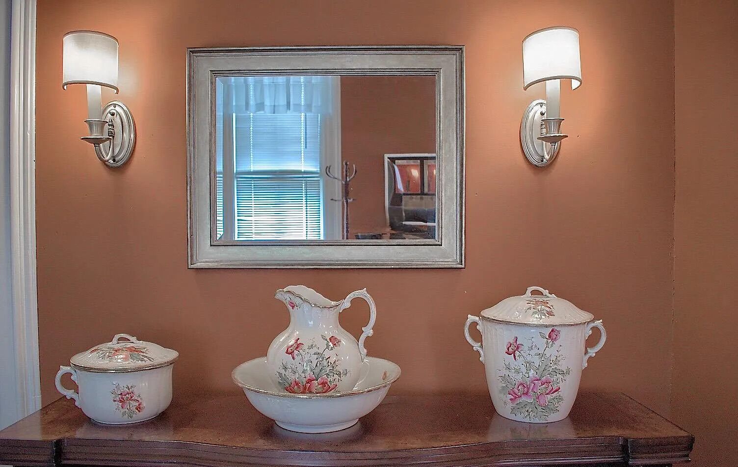 Un tocador de baño con espejo, dos apliques de pared y tres adornos florales de cerámica.