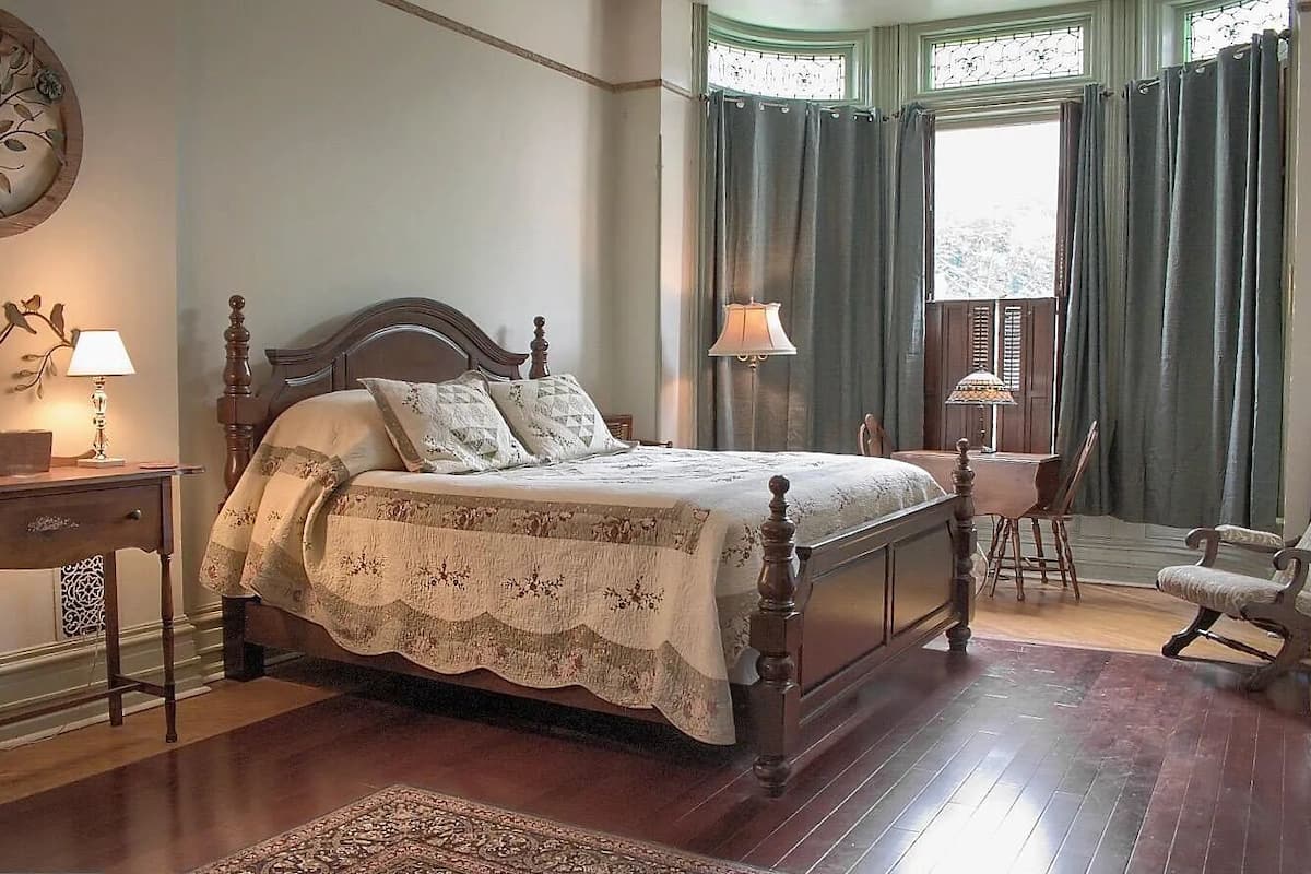 A traditional bedroom with a four-poster bed, a desk with a lamp, a chair, and a window with curtains.