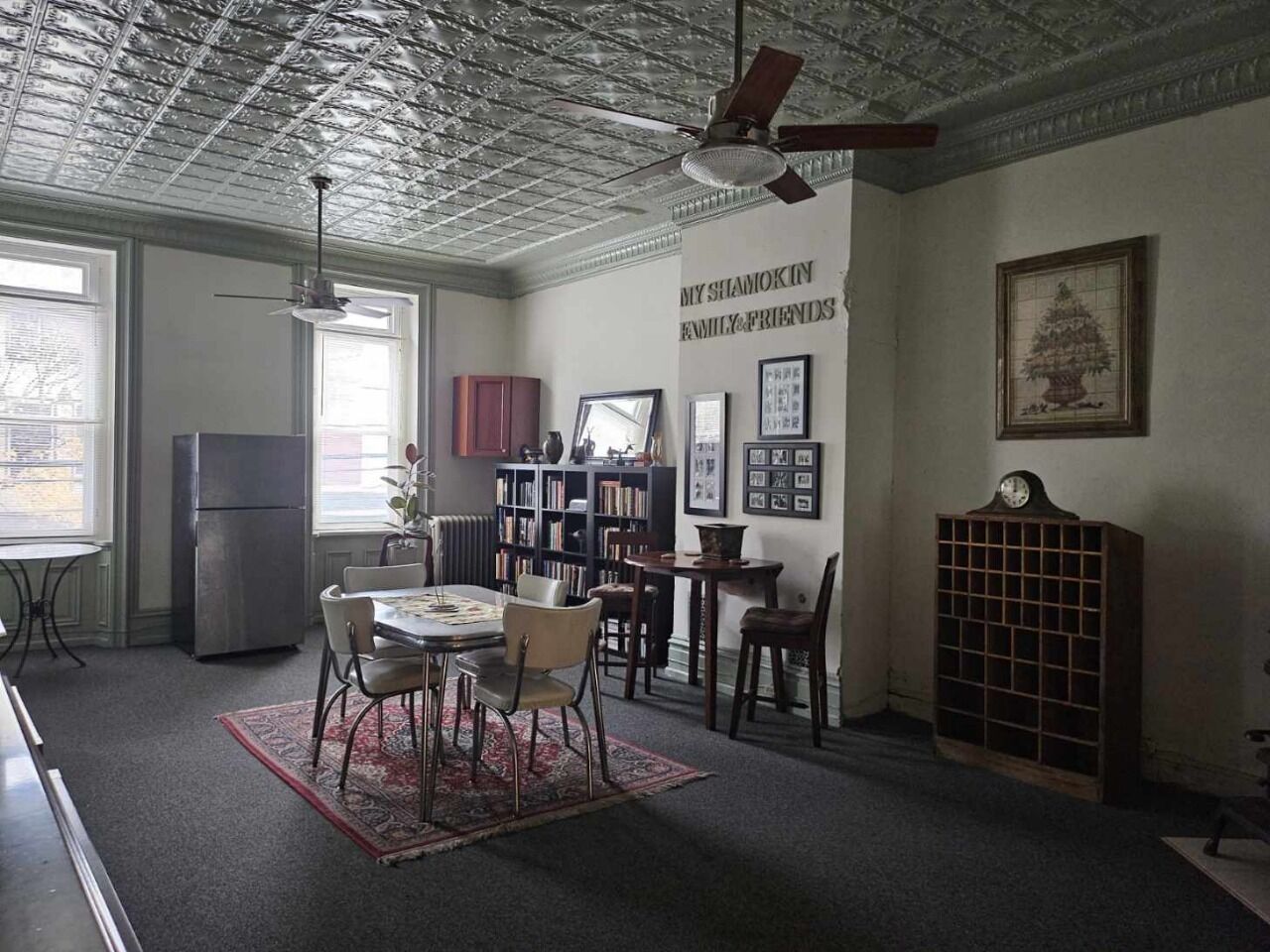 A room with a dining table, chairs, a bookshelf, a radiator, and a clock on the wall.