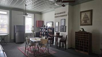 A room with a dining table, chairs, a bookshelf, a radiator, and a clock on the wall.