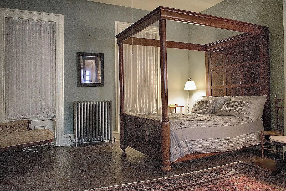 A four-poster bed with a wooden frame, a radiator, a sofa, and a lamp on a nightstand.