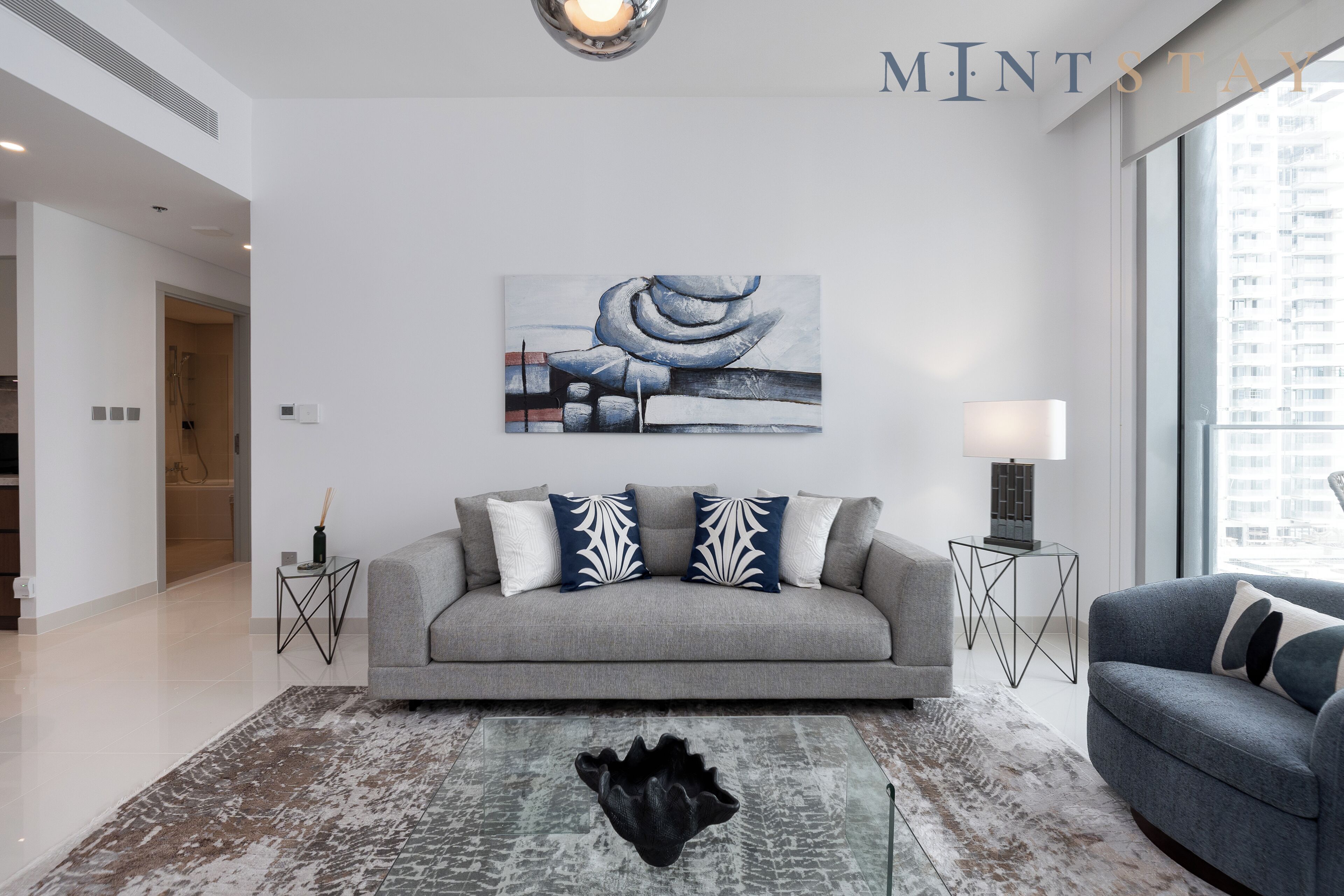 A modern living room with a grey sofa, decorative pillows, a black abstract rug, and a cityscape painting on the wall.