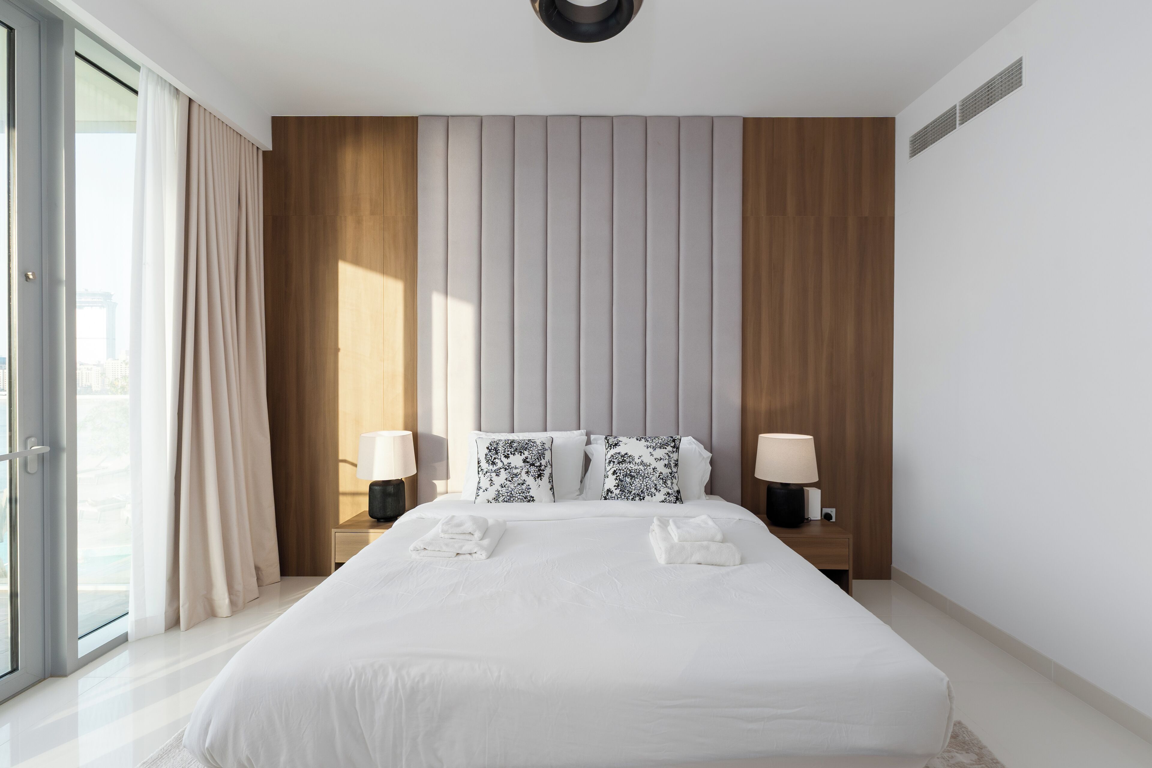 A modern hotel room with a large bed, two bedside tables with lamps, a wooden headboard, and a large window with curtains.